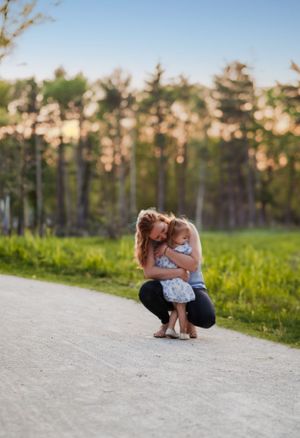A tender moment between parent and child crouching together on a dirt path with evergreen trees and sunset in background.