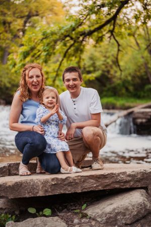 A happy family poses together on large rocks by a stream in a lush green forest setting on a warm summer day.