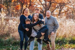 A group of people laugh and embrace while posing outdoors among autumn foliage and tall grass at sunset.