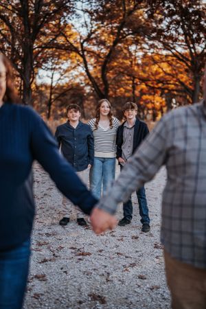 Family group photo in autumn forest with warm fall colors and gravel path, framed by couple holding hands in foreground.