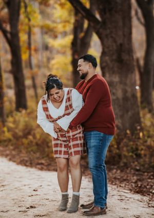 A couple laughs together on a path surrounded by autumn trees with golden fall foliage in the background.
