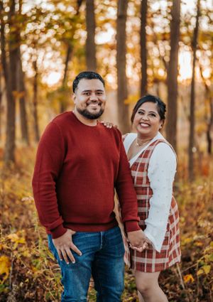A couple poses together in autumn woods during golden hour sunset, wearing red and white casual attire.