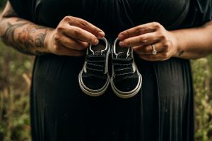 Tattooed hands hold tiny black baby shoes against a dark background in an intimate pregnancy announcement photo.