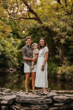 A family poses together on rocky ledge by a peaceful pond surrounded by autumn trees in golden light.
