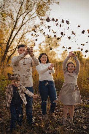 A family enjoys an autumn day outdoors throwing fall leaves in the air during a golden sunset photoshoot.