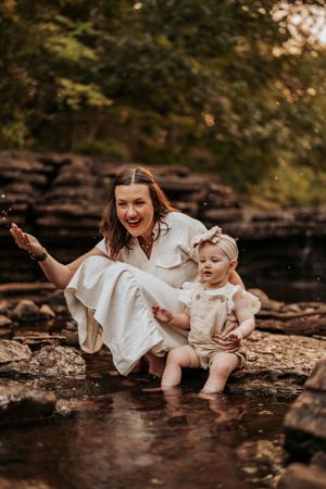 Mother and baby in white outfits share a joyful moment playing at the edge of a creek in a wooded setting.