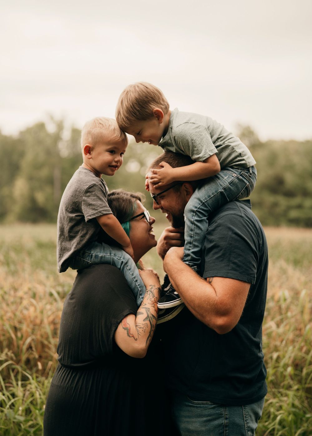 Candid family portrait of parents holding two young children on shoulders in a golden field at sunset.