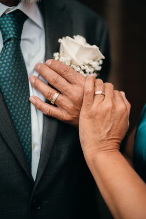 Close-up of wedding rings and white rose boutonniere on a groom wearing a dark suit and teal patterned tie.
