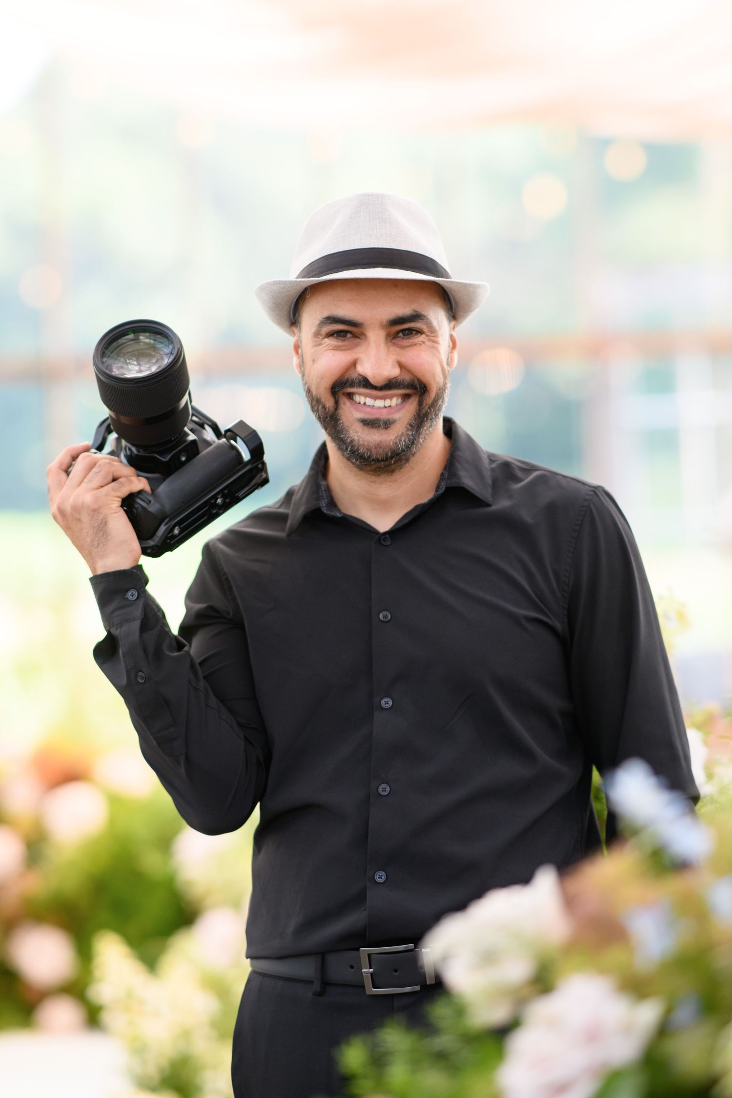 Professional photographer in black shirt and white hat holding camera equipment in outdoor setting.