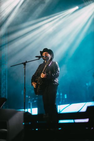 Country musician performing on stage with guitar and cowboy hat under dramatic blue spotlights.