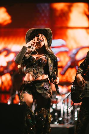 Female performer in cowboy hat and embellished western outfit singing on stage with dramatic fiery backdrop.