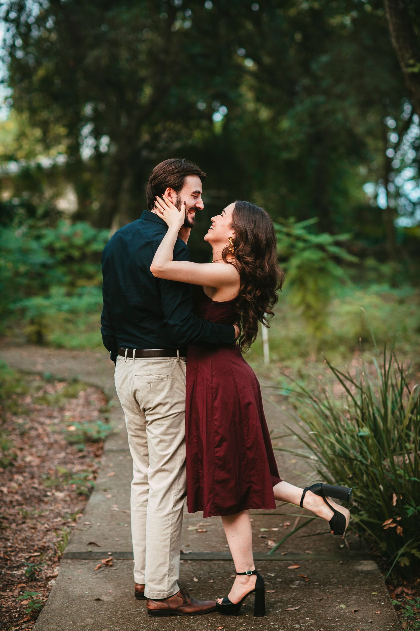 A couple embraces and laughs together on a garden path, woman in burgundy skirt and black top, man in dark shirt and khakis.