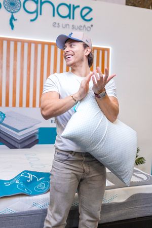 Person in baseball cap and white t-shirt laughing while holding a pillow in front of mattress display at bedding store.