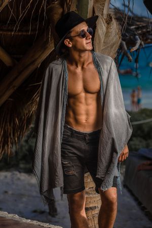 Muscular man in sunglasses and open grey shirt posing against tropical palm tree backdrop on sunny beach.