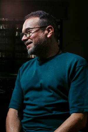 A person in a teal sweater smiles warmly in a dimly lit setting.