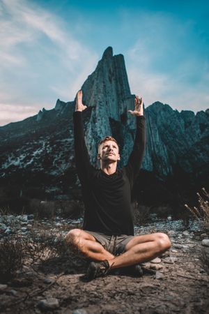 Person meditating in lotus pose with raised arms against dramatic mountain landscape at sunset.