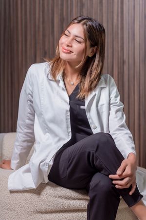 Healthcare professional in white lab coat and dark scrubs relaxes on beige chair against striped wall background.