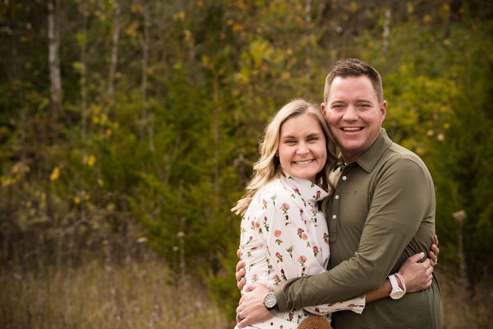 Happy couple embracing outdoors in autumn forest setting, smiling for engagement photo among colorful fall foliage.