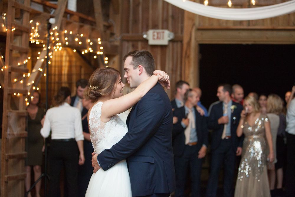 Bride and groom share first dance in rustic barn venue with string lights and guests watching in background.