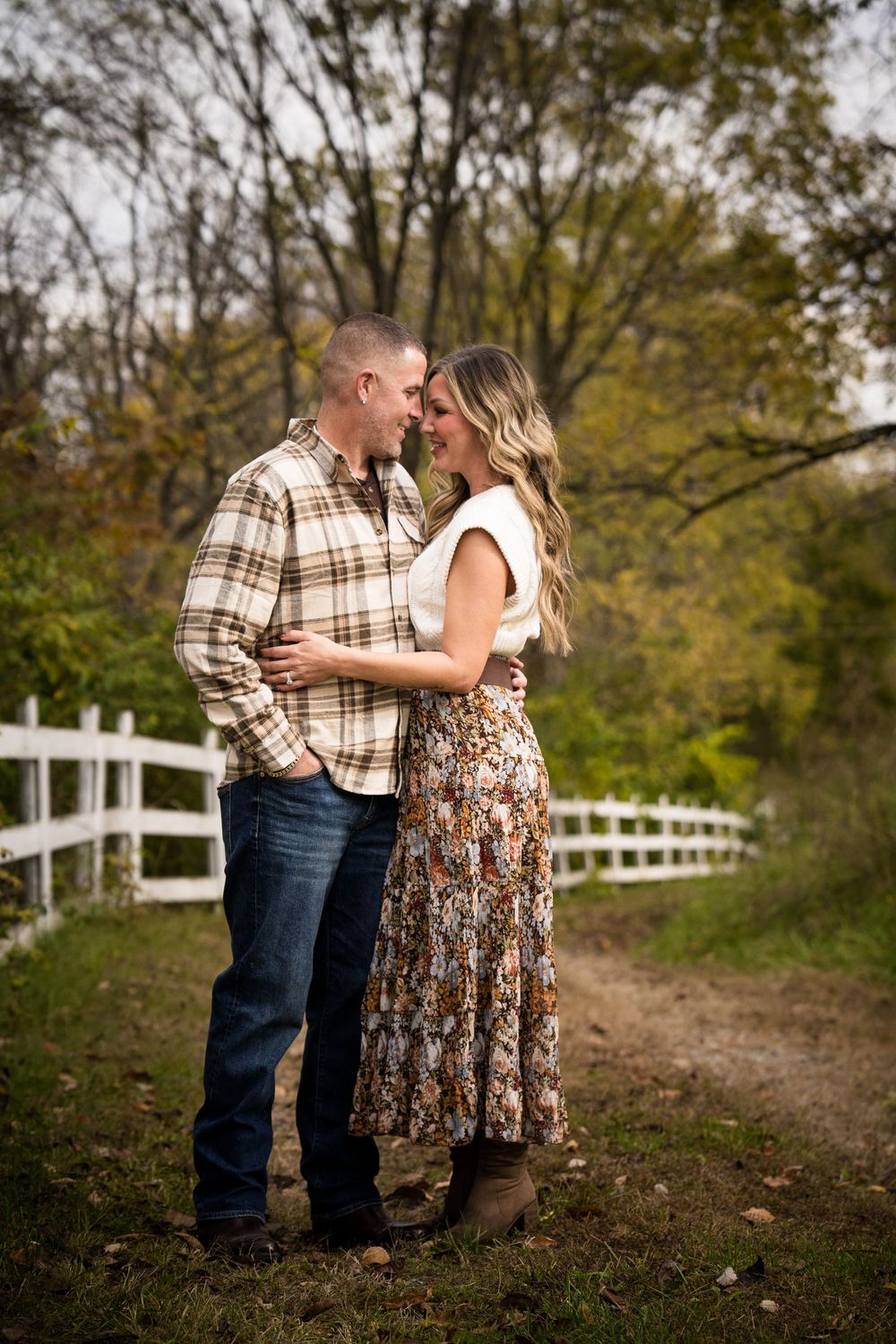 Couple embracing on a rustic dirt path beside a white fence surrounded by autumn trees during a fall engagement photo session.