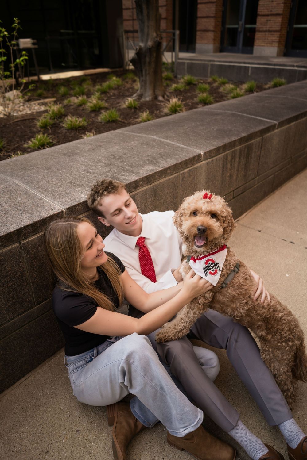 A couple sits outdoors with their fluffy goldendoodle wearing a red bandana, sharing a joyful moment on concrete steps.