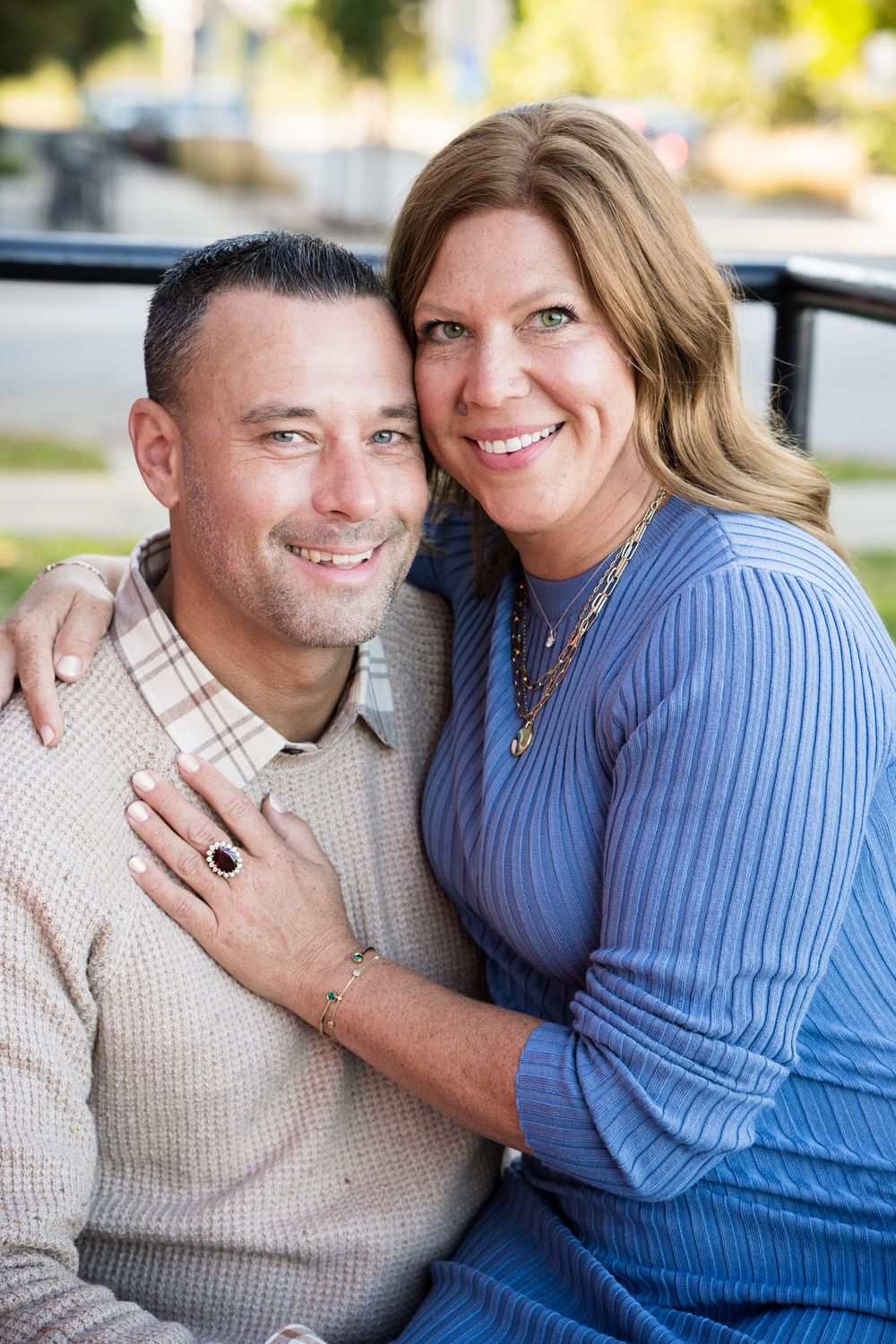Smiling couple posing outdoors, woman in blue top embracing man in plaid shirt, both wearing jewelry.