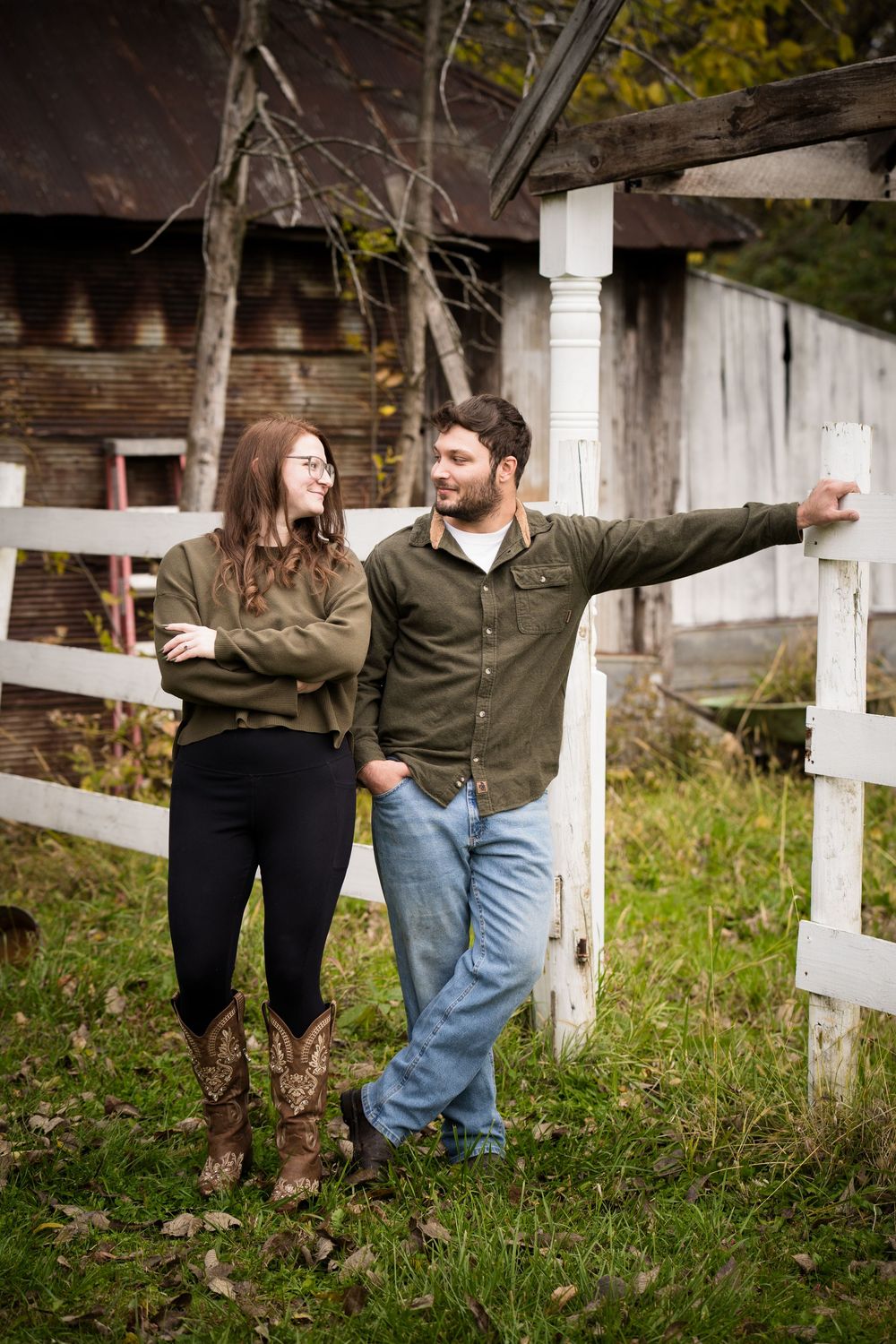 A young couple in matching olive green shirts stand by a white fence near a rustic barn in autumn.