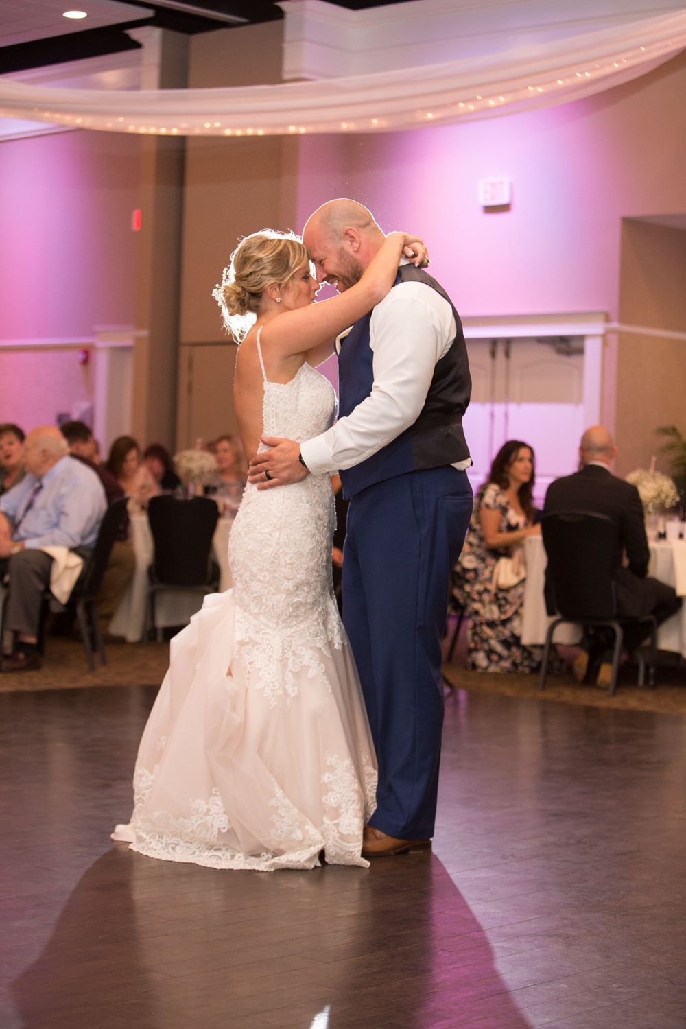 Bride and groom share their first dance at a wedding reception with pink ambient lighting and guests seated in background.