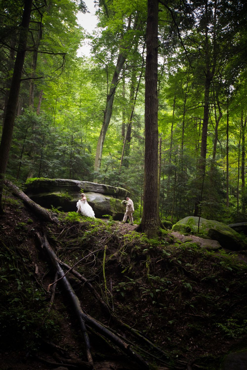 Bride and groom walking through a lush green forest with moss-covered rocks during an outdoor wedding.