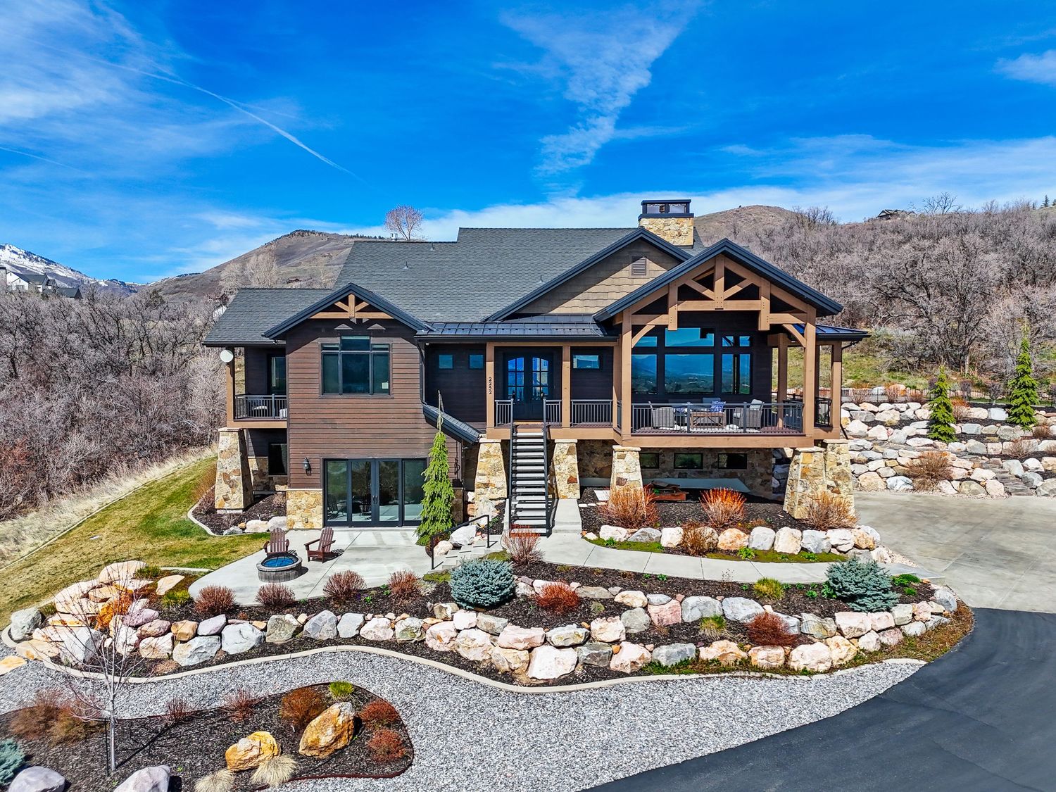 Luxury mountain home with stone landscaping, wood beam accents, large windows, and scenic views under a clear blue sky.