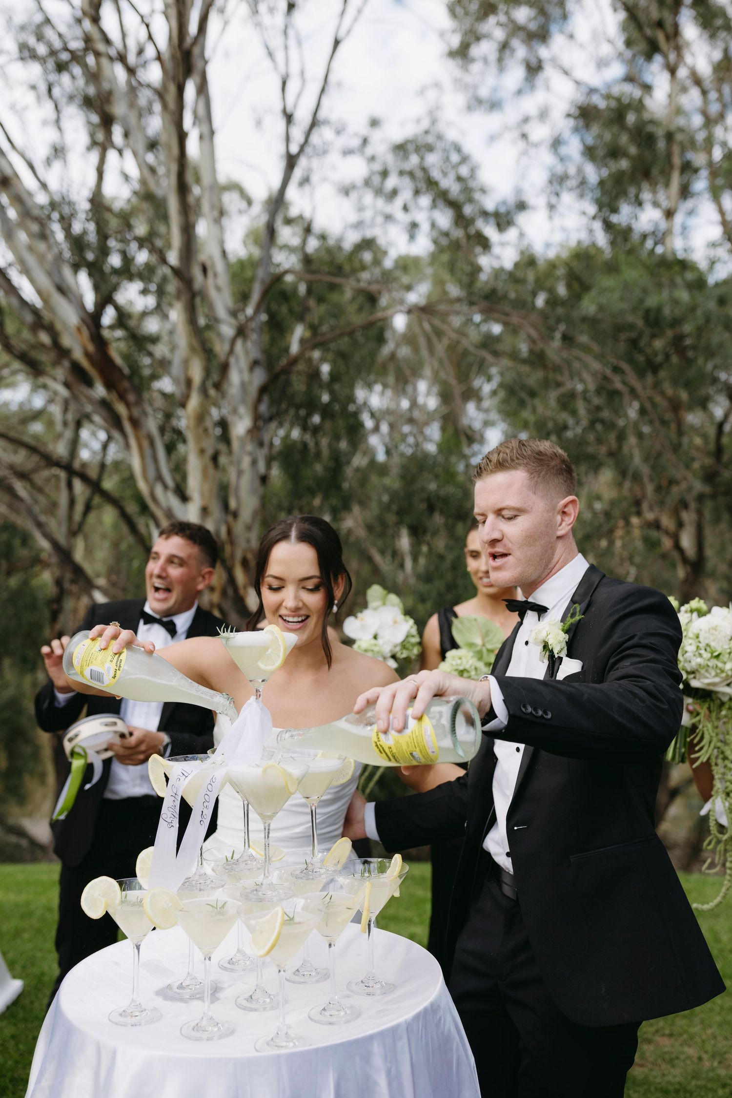 A bride and groom pour champagne into a tower of glasses outdoors, surrounded by their wedding party.