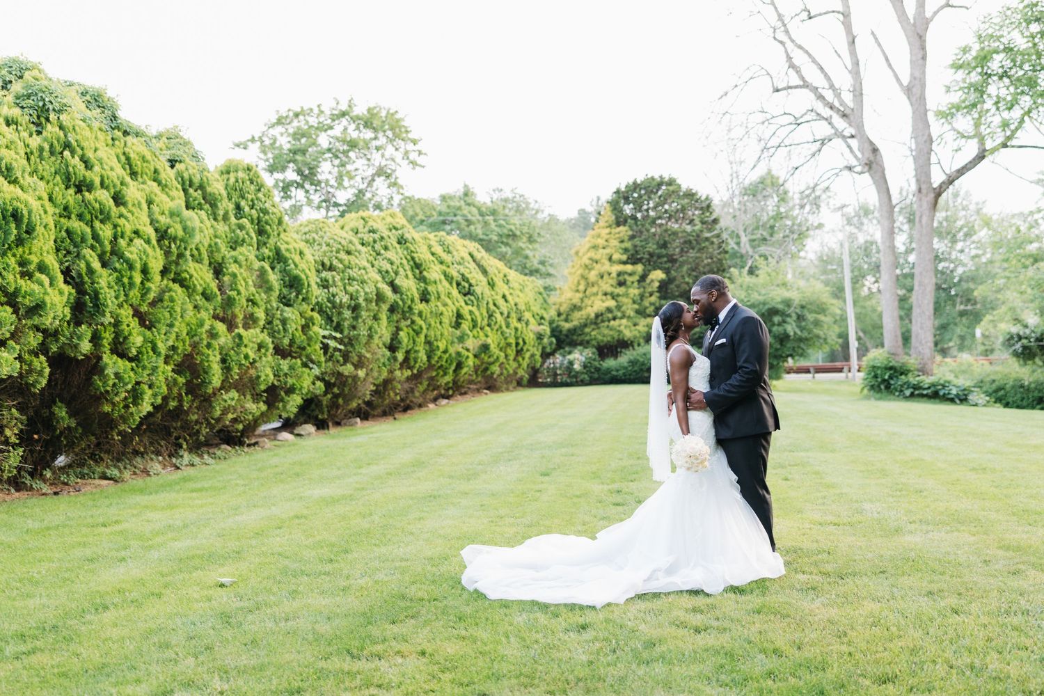 A bride and groom share a romantic kiss on a lush green lawn surrounded by trees on their wedding day.