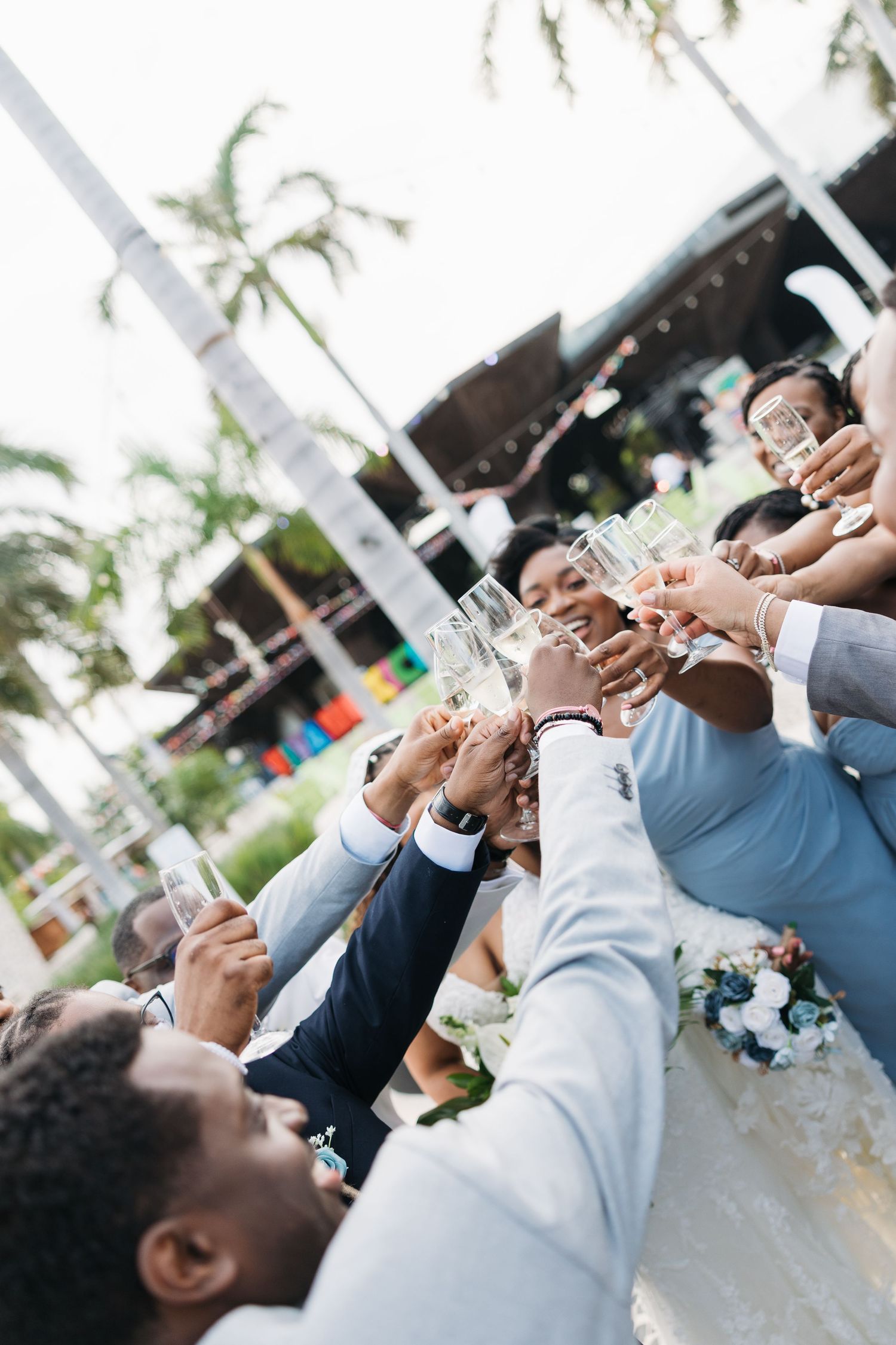 Wedding party clinking champagne glasses outdoors with palm trees and tropical venue in background.