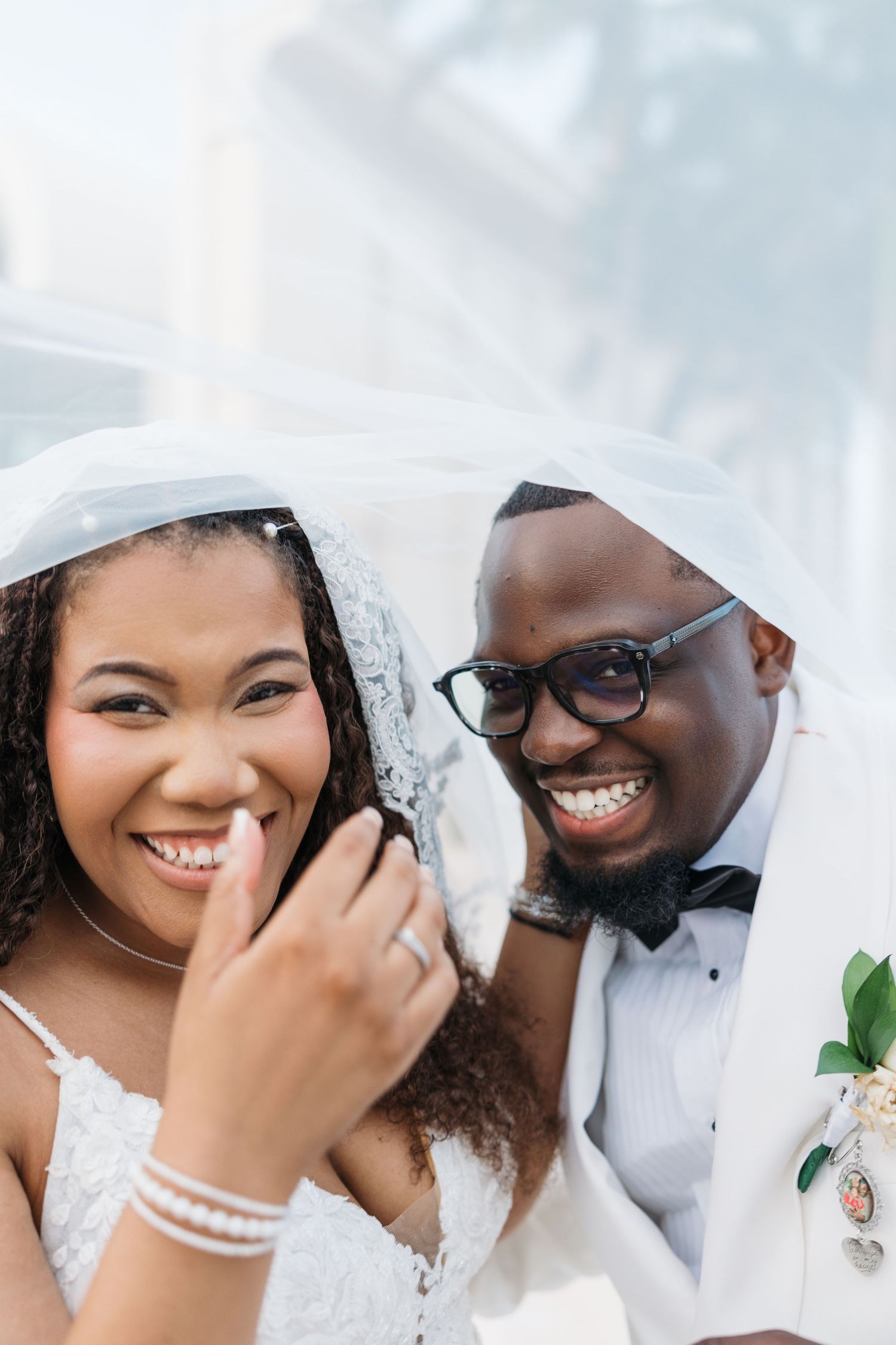 Joyful Black bride and groom smiling under wedding veil, groom in white tuxedo with boutonniere and glasses.