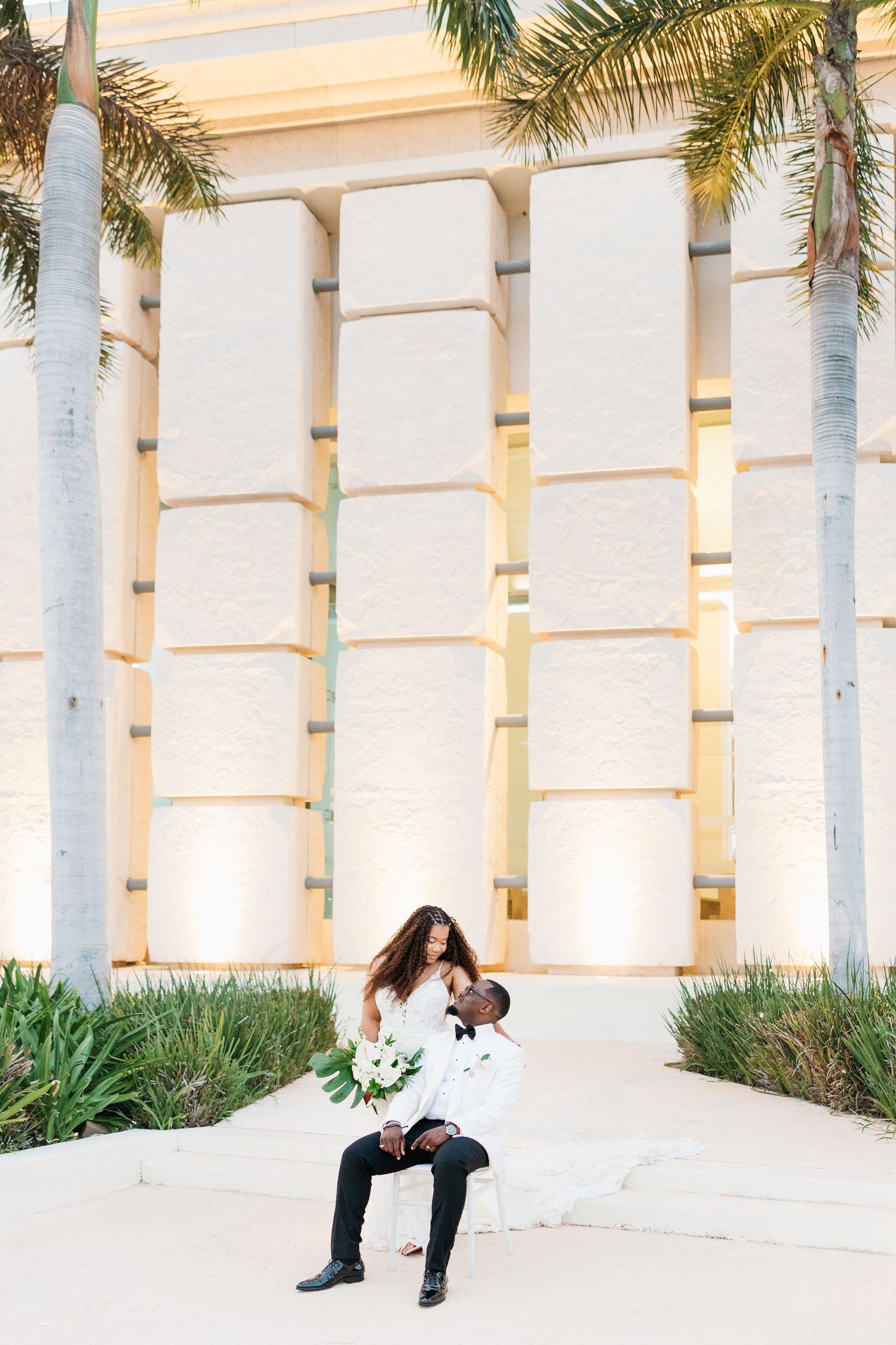 Groom holds bride on his lap outdoors near illuminated columns and palm trees in elegant wedding attire.