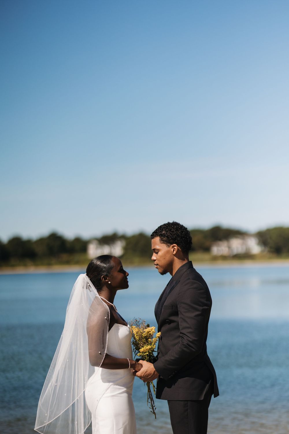 A couple shares a romantic moment by a lake on their wedding day, with the bride in white and groom in black suit.