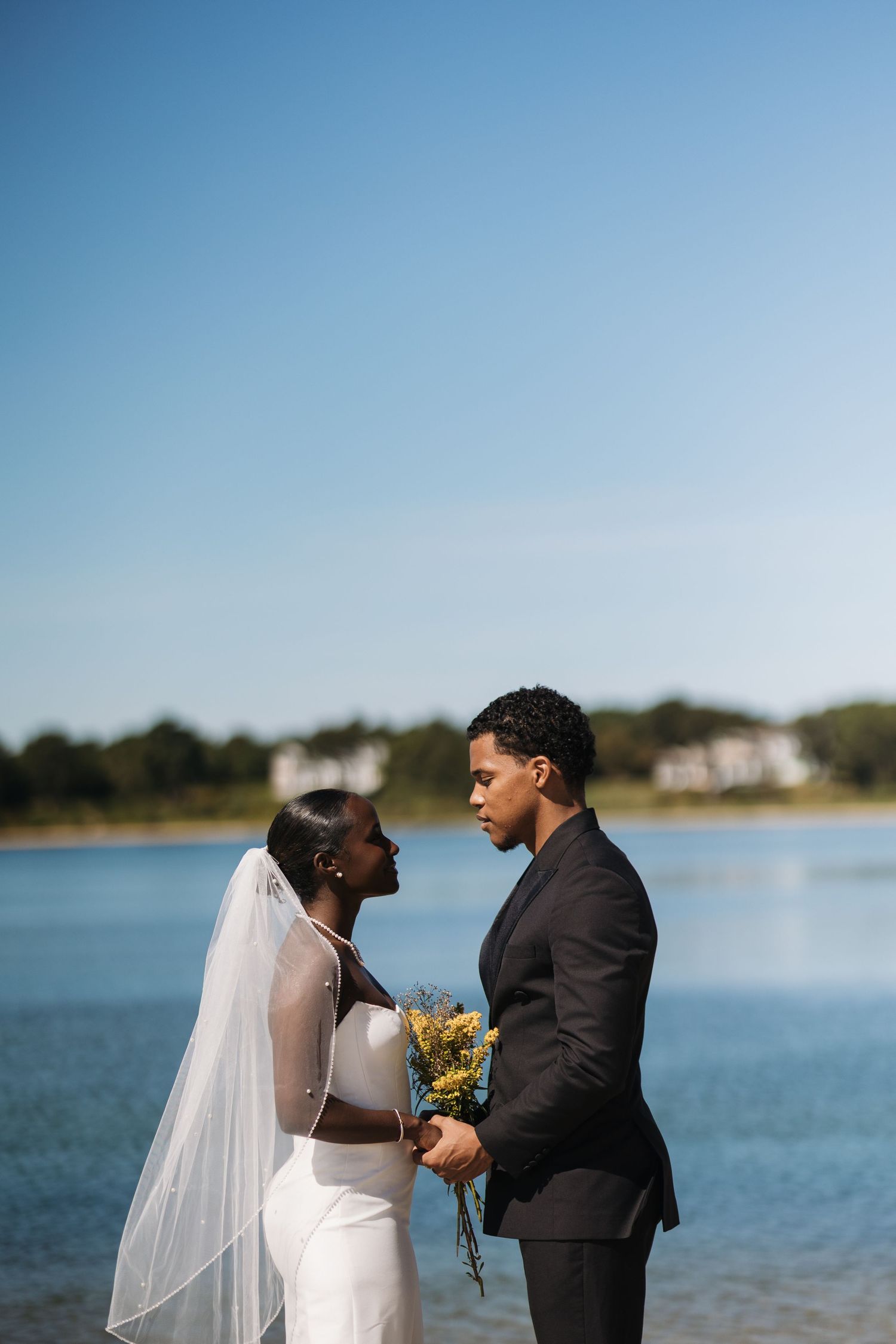 A couple shares a romantic moment by a lake on their wedding day, with the bride in white and groom in black suit.