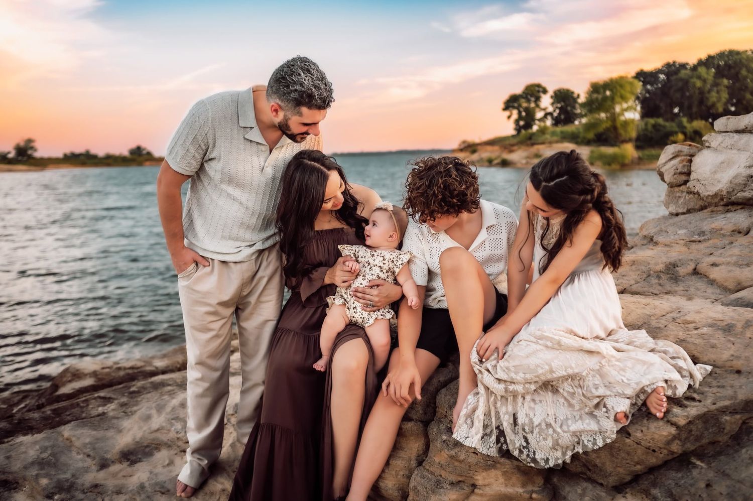 Family of five gathered on rocky shore at sunset, parents and children bonding over adorable baby by a lake.