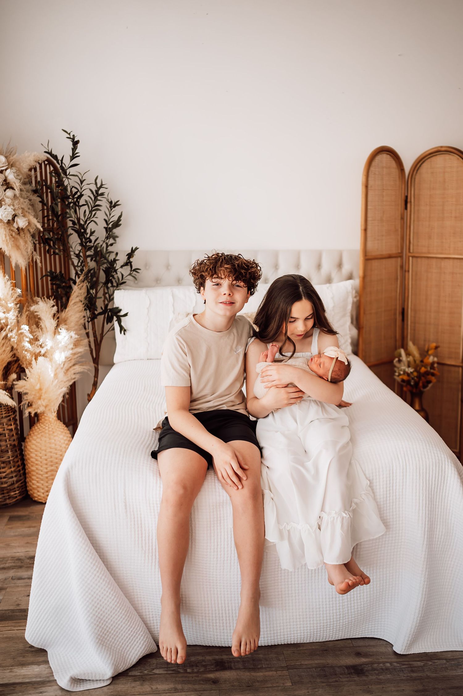 Teen boy and girl sitting on white bed holding newborn baby in boho bedroom with rattan decor.