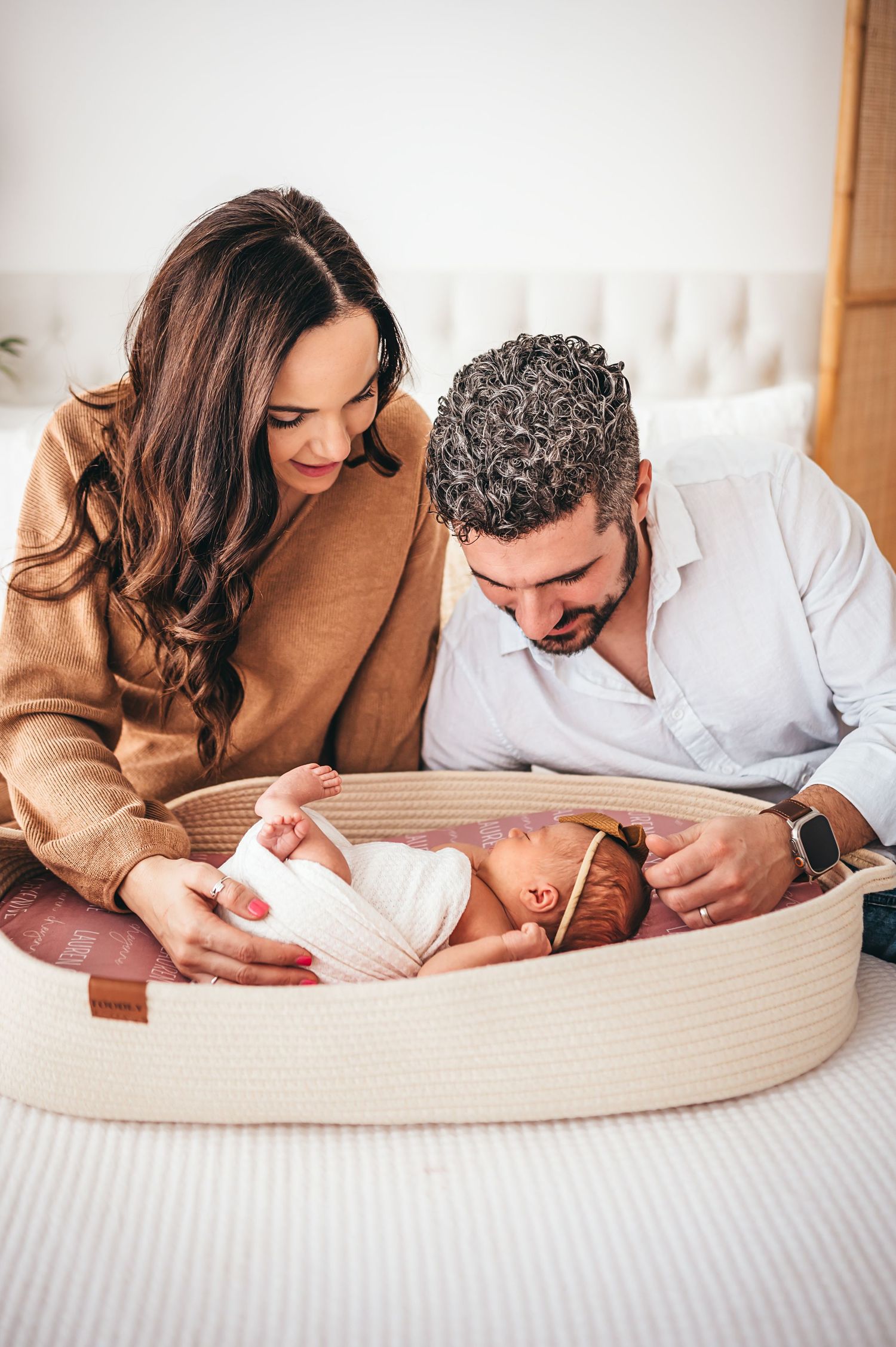 Parents lying on bed gazing lovingly at swaddled newborn baby in woven Moses basket.