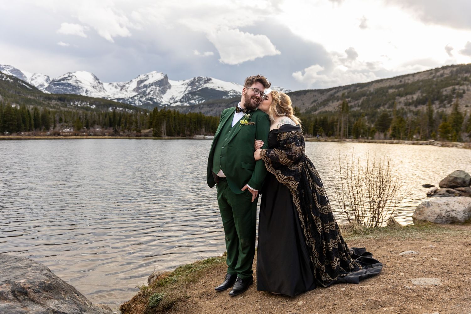 Bride and groom in dramatic black and gold wedding attire posing by a mountain lake with snowy peaks.