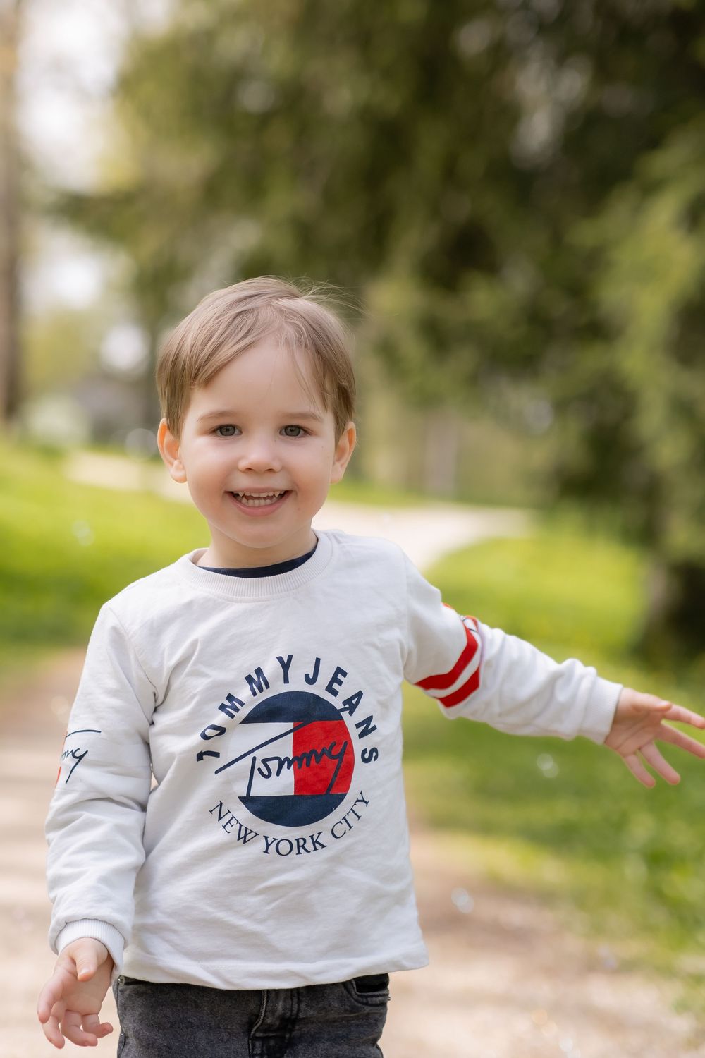 A young child in a white Tommy Jeans sweatshirt smiles while walking outdoors on a sunny day.