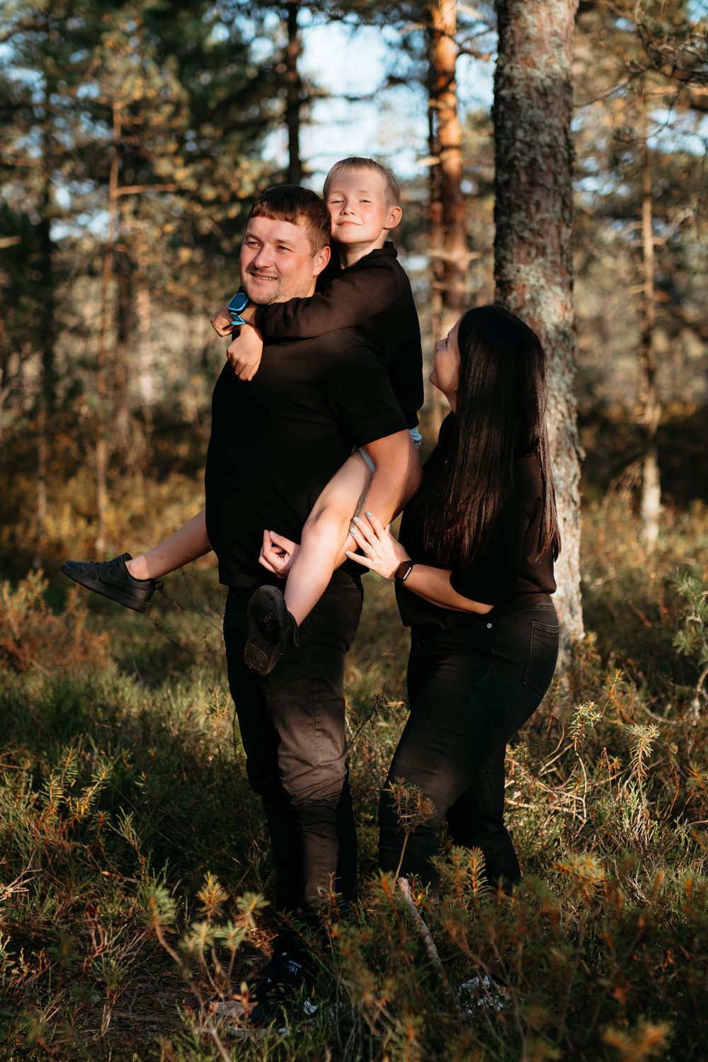 A family wearing black outfits poses together in a sunlit forest with pine trees in the background.