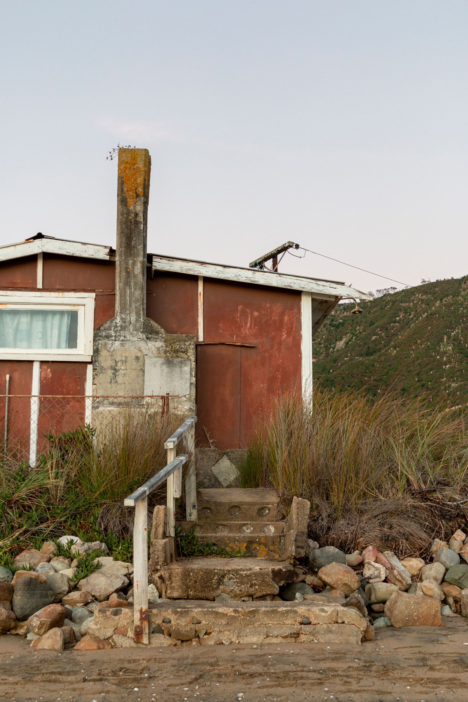 Weathered red cabin with a stone chimney and overgrown steps, set against rocky ground and hills under a pale sky.