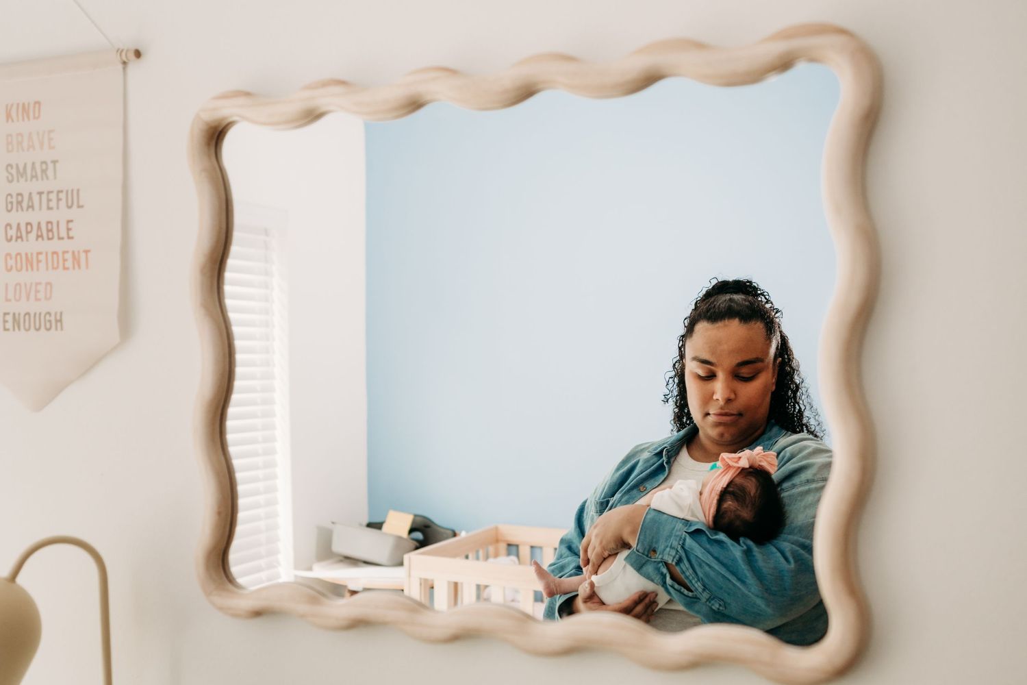 Mirror reflection shows new mom in a blue sweater  holding a baby in a cozy nursery during in home lifestyle newborn photo shoot in Tacoma.