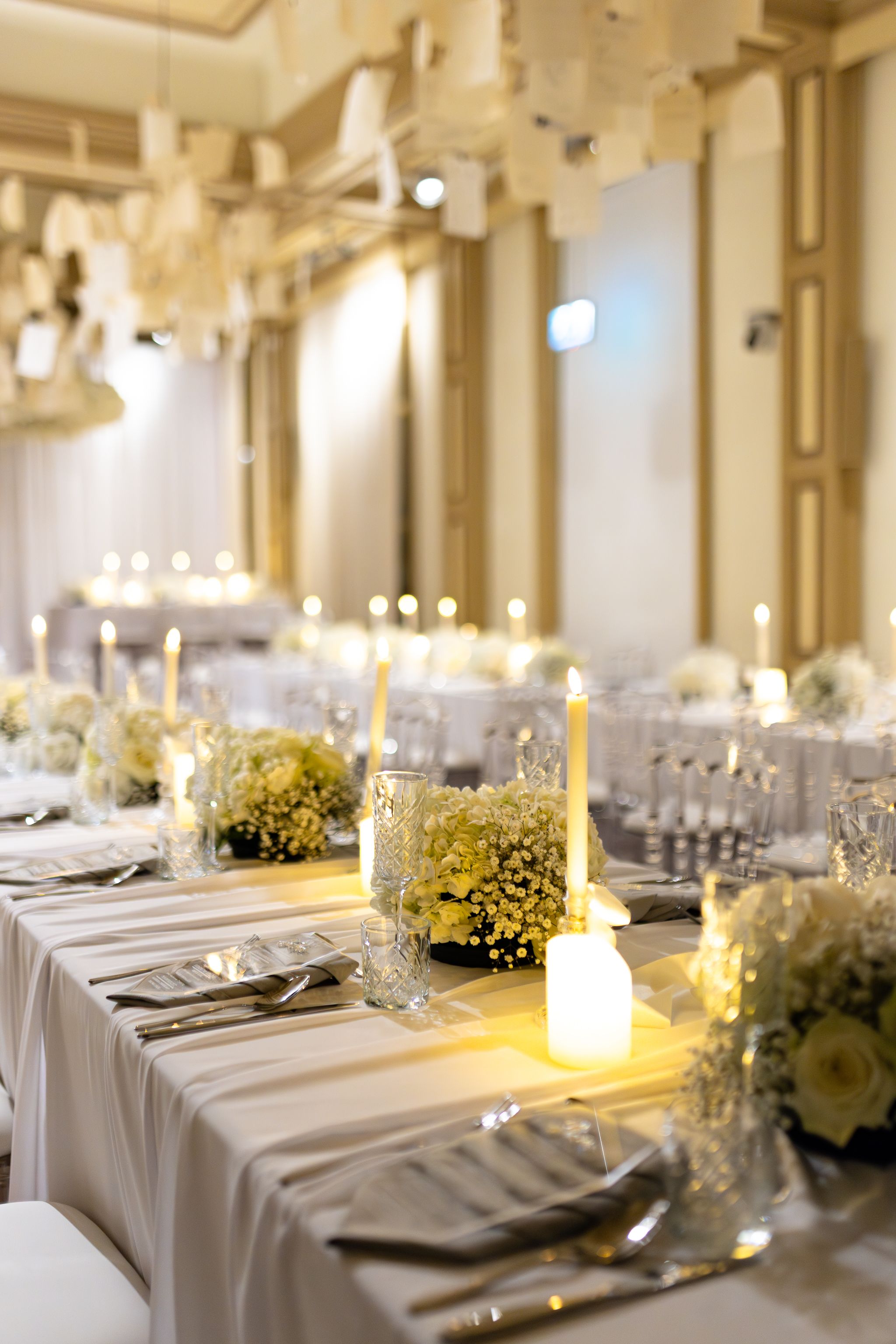 Elegant wedding reception tables set with white linens, glowing candles, and floral centerpieces in a grand ballroom.