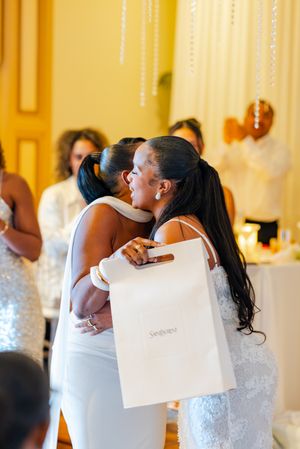 Wedding guests in white dresses embracing while holding shopping bags at an elegant reception.