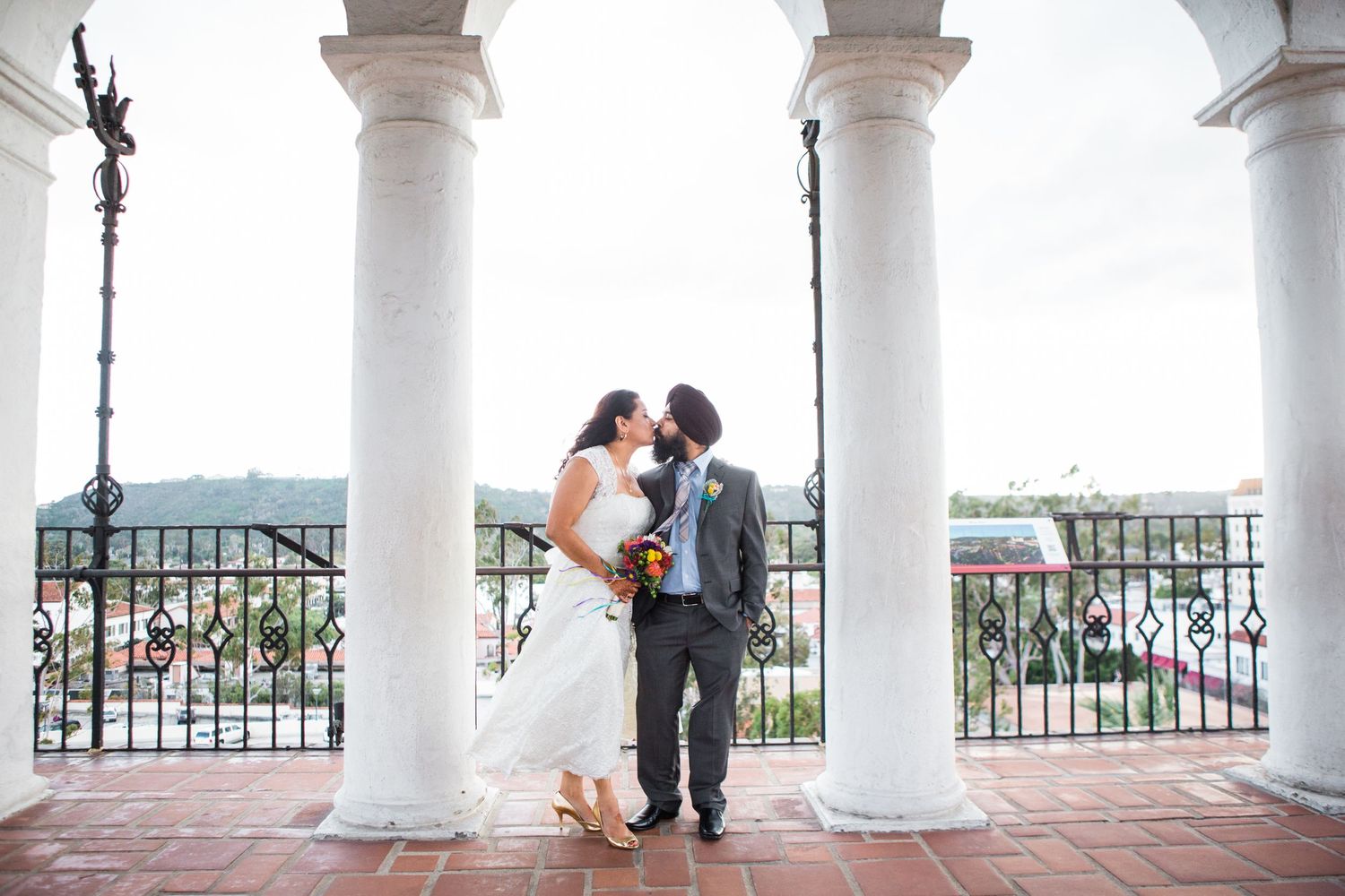 A couple shares an intimate moment under white pillars on a balcony overlooking a scenic view.