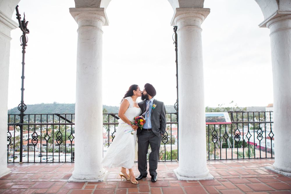 A couple shares an intimate moment under white pillars on a balcony overlooking a scenic view.