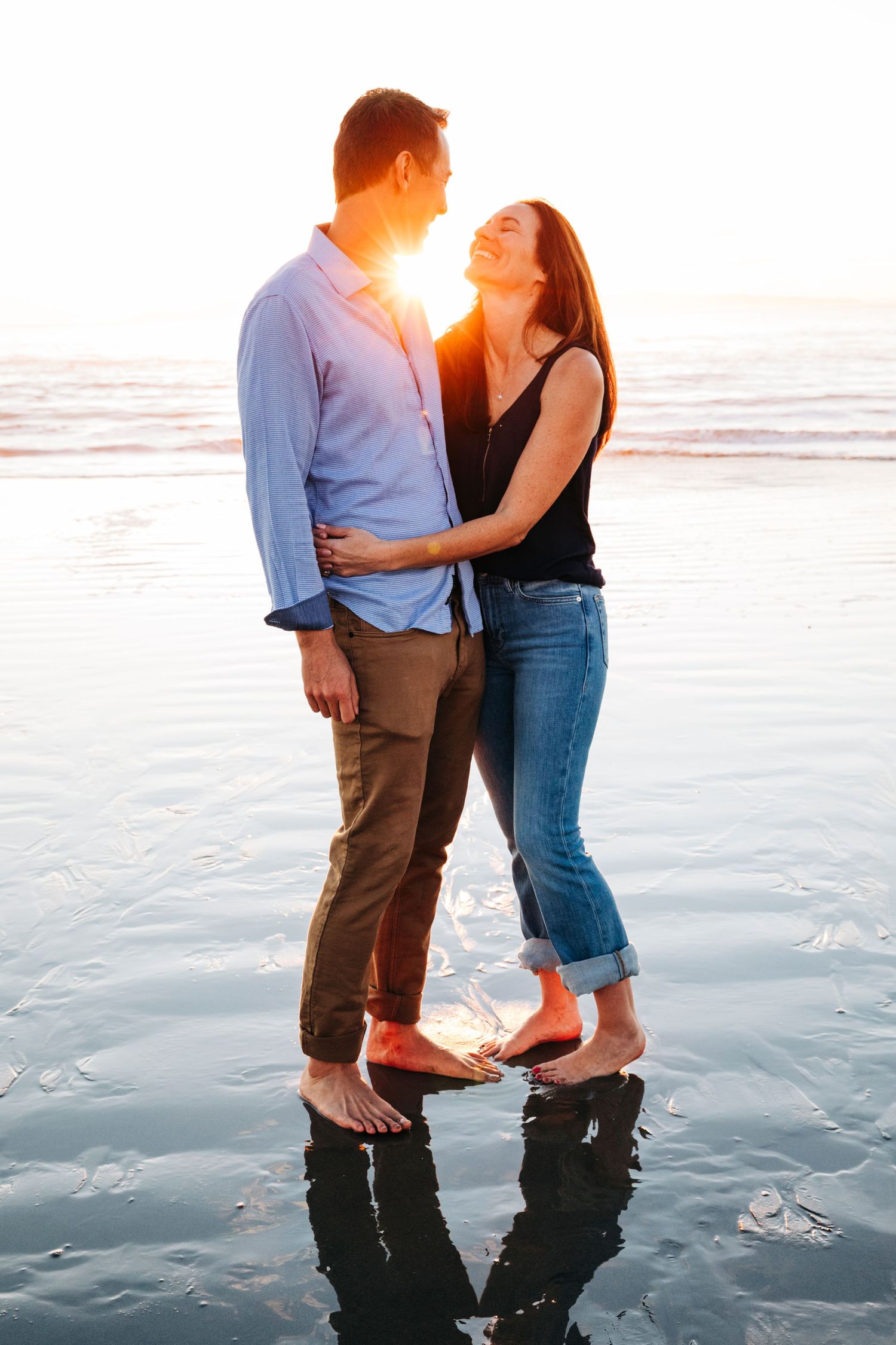 A romantic couple embraces on the beach at sunset with their reflection visible in the wet sand.